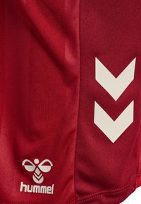 Röda sportshorts i slätt tyg, med vita chevronränder och en logotyp i det nedre hörnet, samt sömnadsdetaljer vid fållen.