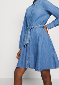 Farkkumekko, jossa on kauluksellinen, napitettava yläosa, pitkät hihat ja vyötäröllä vyö. Mekossa on liehuva hame, keskisininen väri ja hienovarainen tekstuuri.