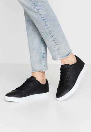 Person iført lyseblå kortere bukser og sorte teksturerede sneakers med hvide såler, stående på en ensfarvet lys overflade.