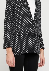 Czarna marynarka w groszki o dopasowanym kroju, z dwoma przednimi kieszeniami, jednorzędowym zapięciem na guzik oraz długimi rękawami z mankietem.