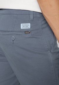 Šedé elastické kalhoty s modro-černou nášivkou značky na zadní kapse a zapínáním na knoflík; mají čistý šev a úzký střih.