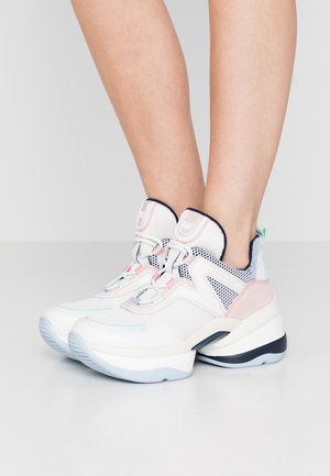 Baskets épaisses blanches et pastel avec des détails en mesh et en cuir, portées pieds nus sur un fond gris.