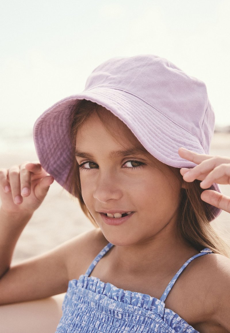 Jeune fille portant un bob violet et une robe fleurie bleue, souriant doucement tout en ajustant son chapeau en extérieur, probablement lors d'une journée ensoleillée.