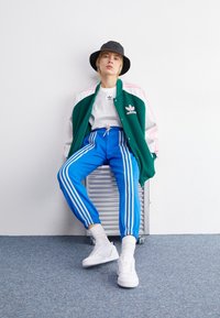 Green and white varsity jacket with pink accents, white t-shirt, blue joggers with white stripes, white sneakers, and a black bucket hat.