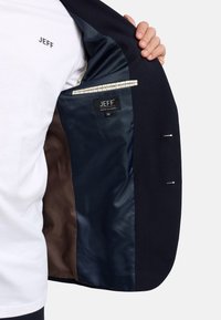 Homme portant une chemise blanche avec le texte "JEFF" tenant un blazer bleu marine ouvert montrant la doublure intérieure bleu foncé et marron ainsi que l'étiquette JEFF à l'intérieur.