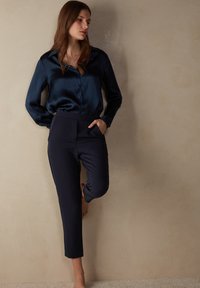 Camicia button-up in satin navy e pantaloni scuri navy su misura con tasche. Texture liscia e design aderente; la camicia ha un colletto classico.