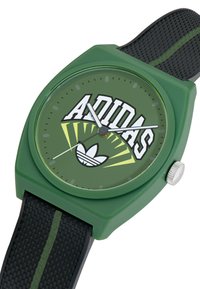 Žalias kvadratinis laikrodis su juodu tekstūruotu dirželiu. Yra ryškus baltas "ADIDAS" logotipas, geltonos detalės ir sidabrinė karūnėlė. Švarus, modernus dizainas.