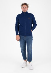 Maglione blu navy con zip e colletto alto, polsini a costine e una piccola toppa con logo, abbinato a jeans azzurri e sneakers bianche.
