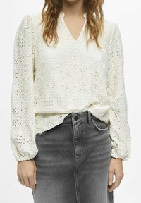 Weißes Spitzenbluse mit V-Ausschnitt, langen Ballonärmeln und kunstvollen Diamantmustern, kombiniert mit einem grauen Jeansrock.