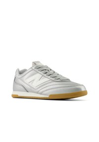 Chaussure de sport argentée avec une tige synthétique lisse, un logo blanc en accent, des lacets gris texturés et une semelle extérieure en caoutchouc gomme.