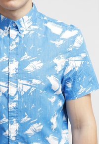 Mężczyzna w niebieskiej koszuli z krótkim rękawem z białymi abstrakcyjnymi wzorami na gładkim jasnym tle.