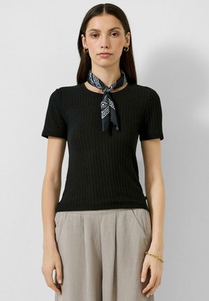 Kobieta ubrana w czarny, teksturowany top z krótkim rękawem, jasnobeżowe spodnie, czarną chustkę na szyję w wzory, złote kolczyki koła oraz złotą bransoletkę.