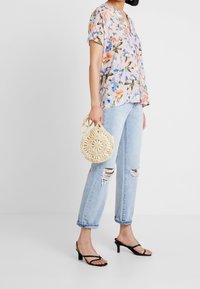 Blomstret kortærmet bluse i pastelfarver, parret med lyseblå slidte jeans og sorte strophæle. Holder en rund, tekstureret cremet taske.