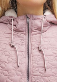 Prošívaná růžová bunda s zapínáním na zip, stahovacími šňůrkami na kapuci a měkkou podšívkou. Má vzor ve tvaru diamantů a kontrastní detaily zipu.