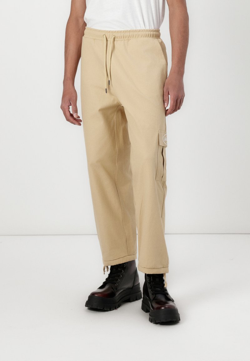 Beige cargo bukser med elastisk talje og snor. Har sidelommer og et broderet logo-patch. Stylet med sorte kampstøvler.