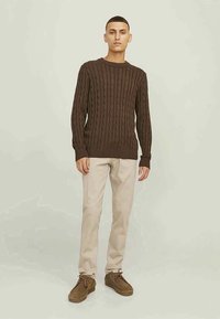 Braun gestrickter Pullover mit gerippten Bündchen, kombiniert mit hellbeigen Hosen und braunen Schuhen. Modell steht vor einem hellen Hintergrund.