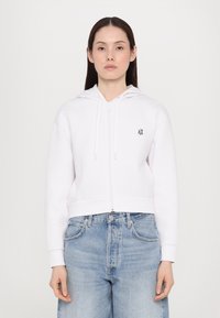 Biela mikina na zips s kapucňou a šnúrkami, so malým čiernym logom na hrudi. Nosená s svetlomodrými džínsami.