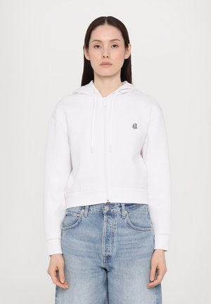 Sudadera blanca con cremallera y capucha, que cuenta con cordones en la parte delantera, y presenta un pequeño logo negro en el pecho. Combinada con unos jeans de mezclilla azul claro.