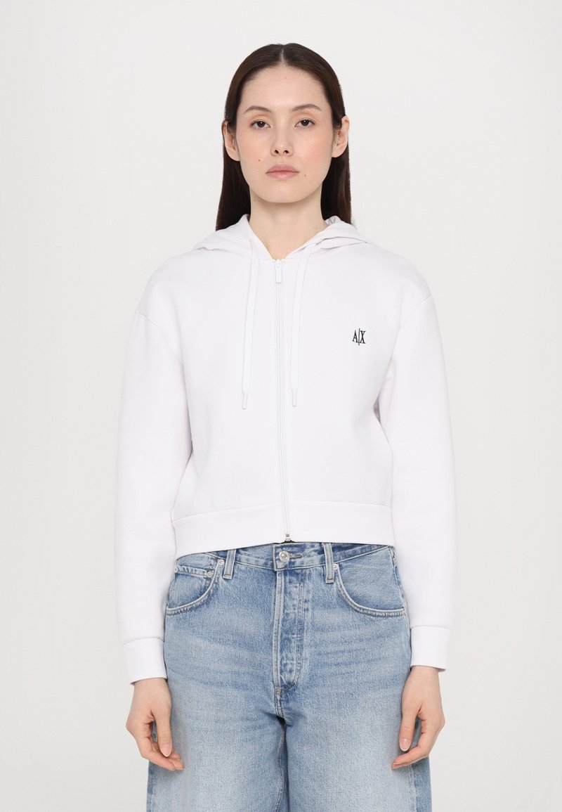 Biela mikina na zips s kapucňou a šnúrkami, so malým čiernym logom na hrudi. Nosená s svetlomodrými džínsami.