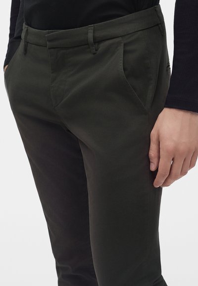 Homme portant un pantalon vert foncé et une chemise noire à manches longues, avec une main dans la poche et l'autre reposant à ses côtés.