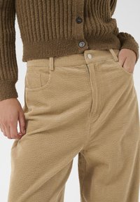 Oseba v bež kordne hlače in rjavem rebrastem puloverju, z eno roko v žepu in drugo sproščeno ob strani.