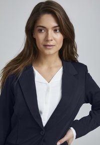 Námornícky blazer s klopovými límci a jedním knoflíkem, vrstvený přes bílou blůzu s V-výstřihem. Hladká textura, přizpůsobený střih.