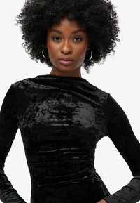 Vestido de terciopelo negro con mangas largas, textura suave, escote alto y silueta ajustada. Presenta un ligero fruncido en la cintura.