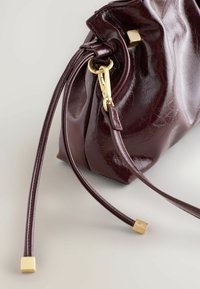 Burgundy nahkakäsilaukku, jossa on poimutettu muotoilu, kultaiset varusteet ja pitkät nahkahihnat, joissa on neliömäiset kultavihjeet. Sujuva pinta.