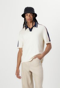 Tricou polo cu mânecă scurtă de culoare crem, cu guler și dungi bleumarin, asortat cu pantaloni bej deschis. Modelul poartă o pălărie neagră tip bucket.