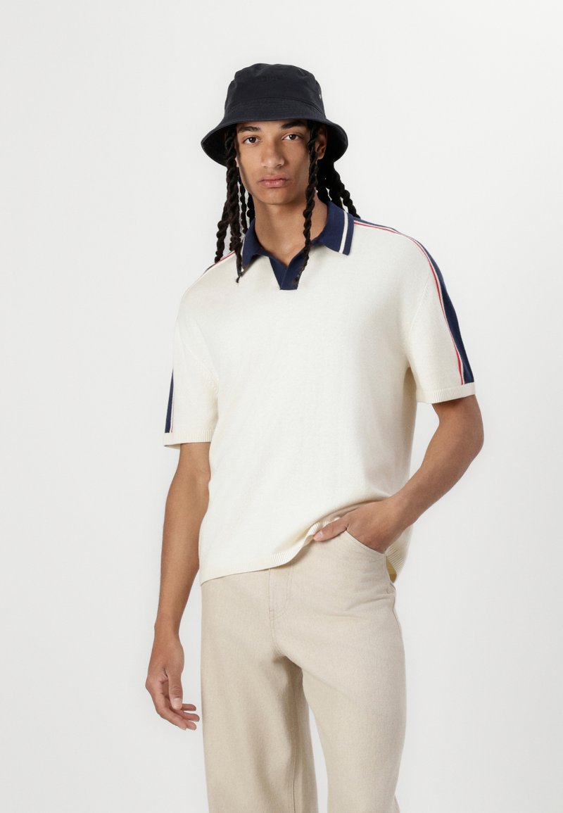 Camisa polo corta de manga con color crema y cuello azul marino con rayas, combinada con pantalones beige claros. El modelo lleva un sombrero de cubo negro.