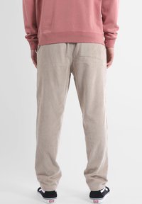 Pantalon en velours côtelé beige à rayures verticales, coupe décontractée, avec taille élastique. Chaussures de sport noires visibles en bas.