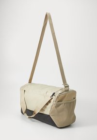 Beige duffelbag met een gestructureerd oppervlak, een zwarte bodem en een lange verstelbare schouderriem. Voorzien van een ritsluiting en een zijzak.