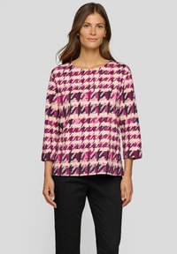 Frau trägt ein langärmliges Oberteil mit rosa, lila und schwarzem Hahnentrittmuster, kombiniert mit schwarzen Hosen, steht vor einem grauen Hintergrund.