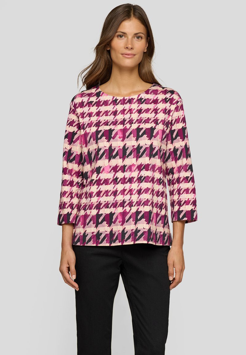 Frau trägt ein langärmliges Oberteil mit rosa, lila und schwarzem Hahnentrittmuster, kombiniert mit schwarzen Hosen, steht vor einem grauen Hintergrund.