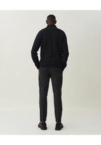 Schwarzes langärmliges Polohemd mit Kragen, kombiniert mit dunkler, strukturierter Hose und schwarzen Turnschuhen, von hinten vor hellem Hintergrund gesehen.