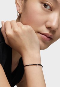Pulsera de cuentas negras con un pequeño encanto de plata, llevada en la muñeca. El modelo tiene un look de maquillaje natural y sostiene su mano cerca de su rostro.