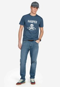 Camiseta azul marino con calavera blanca y texto "HARPER", combinada con jeans azul claro y zapatillas multicolores. Una gorra gris completa el atuendo.