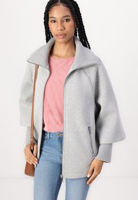 Grå oversized zip-jakke med høy krage, ribbede mansjett-detaljer og sidelommer, båret over en rødstripet topp og blå jeans.
