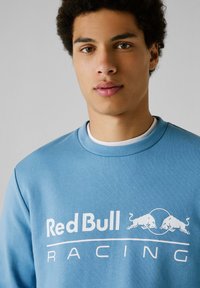 Svetlo modra dukserica s slikovnim logotipom "Red Bull Racing" v beli barvi, z dvema grafičnima bikoma in okroglo potezo pod besedilom. Gladek flisasti material.