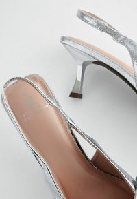 Hopeakylkiset slingback-korkokengät, joissa on kiiltävä pinta, avonaiset varpaat ja kapeneva, matala korko. Sisäpohja on vaaleanbeige.