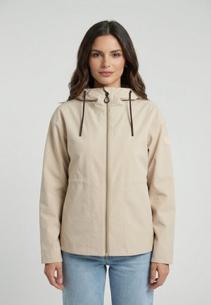 Femme aux cheveux bruns ondulés portant une veste à capuche beige zippée et un jean bleu clair, debout devant un fond clair uni.