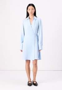 Abito avvolgente azzurro chiaro con maniche lunghe, tessuto testurizzato, colletto, vita con lacci e orlo a lungo fino al ginocchio. Indossato con ballerine nere con decorazioni.