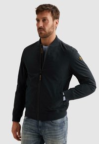 Muž s krátkymi hnedými vlasmi a bradou, oblečený v čiernej bunde na zips a modrých džínsoch, stojaci s jednou rukou v vrecku, pozerajúci sa doľava.