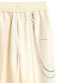 Pantalones de chándal color crema con cintura elástica, que cuentan con bolsillos frontales y un accesorio de cadena de plata adjunto a la cadera.