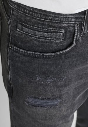 Vaqueros de mezclilla gris oscuro con un pequeño bolsillo para monedas encima de un parche desgastado en el muslo derecho, llevados con una camisa blanca metida por dentro.