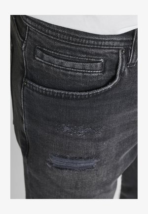 Dunkelgraue Denim-Jeans mit einer kleinen Münztasche über einem verwaschenen Fleck am rechten Oberschenkel, kombiniert mit einem weiß eingesteckten Hemd.