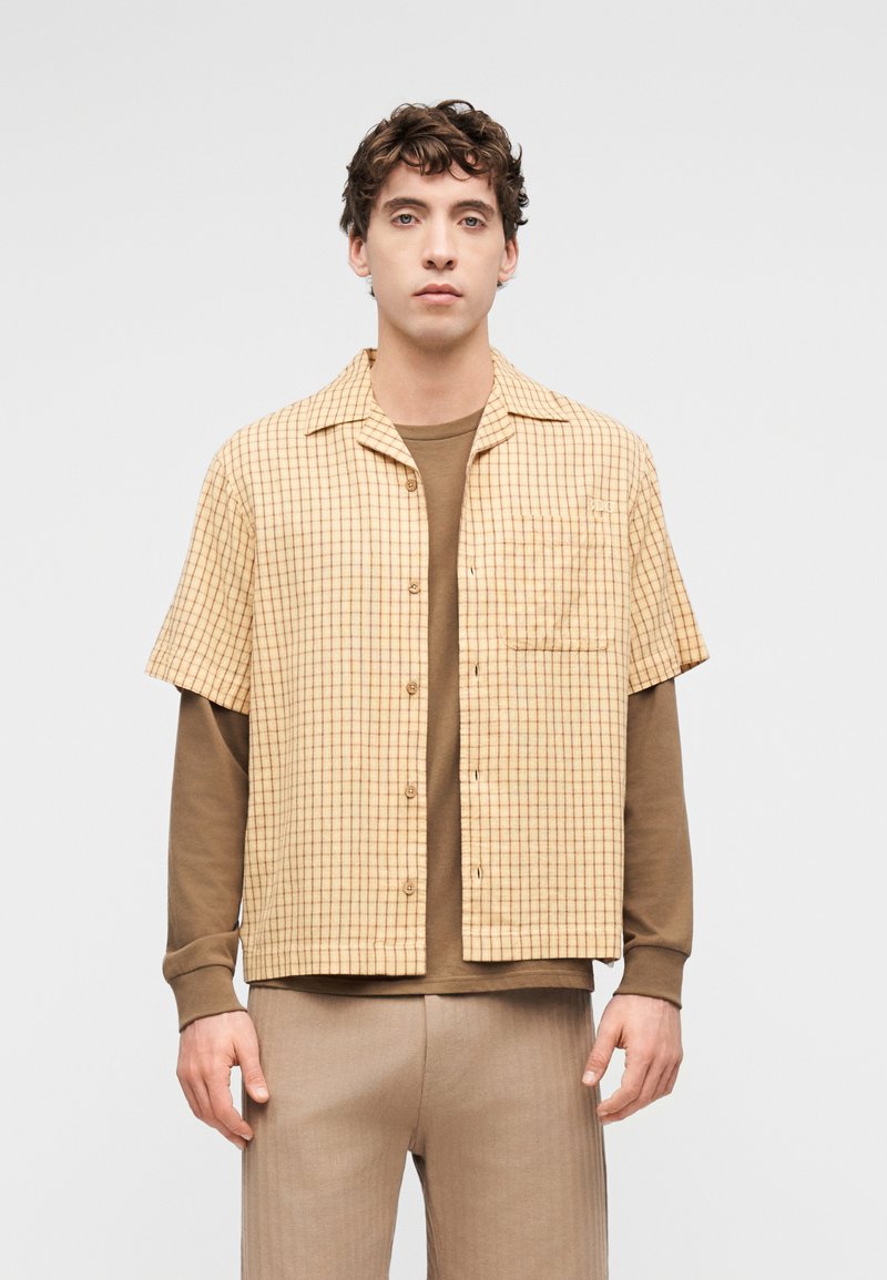 Νεαρός άνδρας που φοράει μπεζ καρό κοντομάνικο πουκάμισο πάνω από καφέ μακρυμάνικη μπλούζα και μπεζ παντελόνι, στέκεται μπροστά σε απλό φωτεινό φόντο.