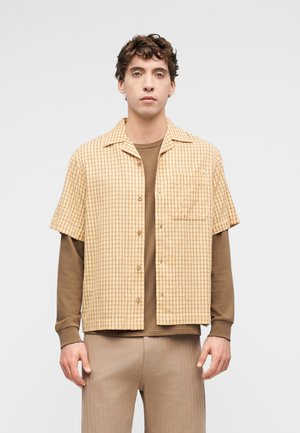 Mladý muž v béžové kostkované krátké košili přes hnědý dlouhý rukáv a béžové kalhoty, stojící před jednoduchým světlým pozadím.