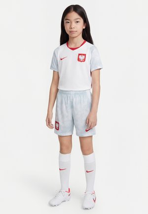 Junges Mädchen steht in polnischer Fußballuniform mit weißem Trikot, hellgrauer Hose, weißen Socken und Fußballschuhen vor einfachem Hintergrund.