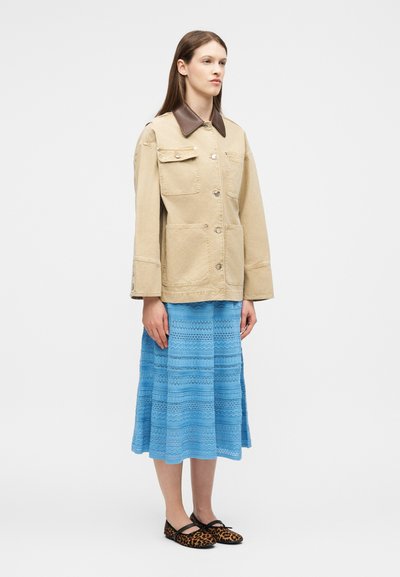 Femme debout, portant une veste beige boutonnée avec un col foncé, une jupe midi bleue à motifs et des ballerines léopard avec des brides.